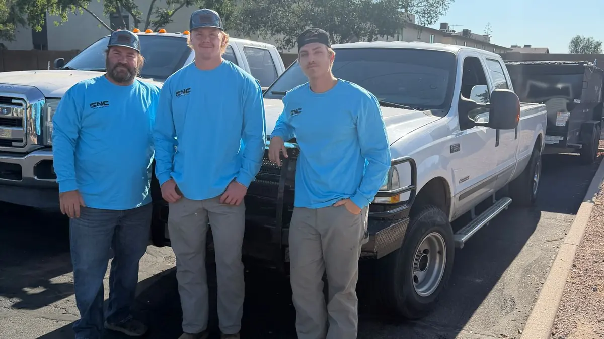 Three CNC Junk Removal crew members in blue branded shirts standing in front of their white pickup truck and dump trailer